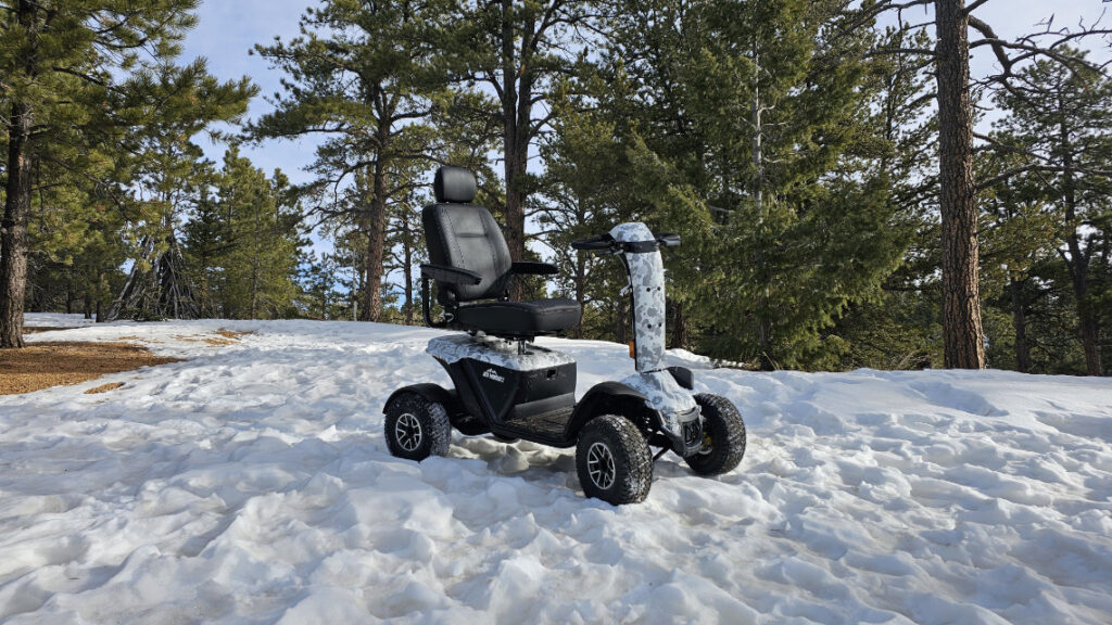 A rugged mobility scooter with large tires is parked on a snowy forest trail surrounded by pine trees.