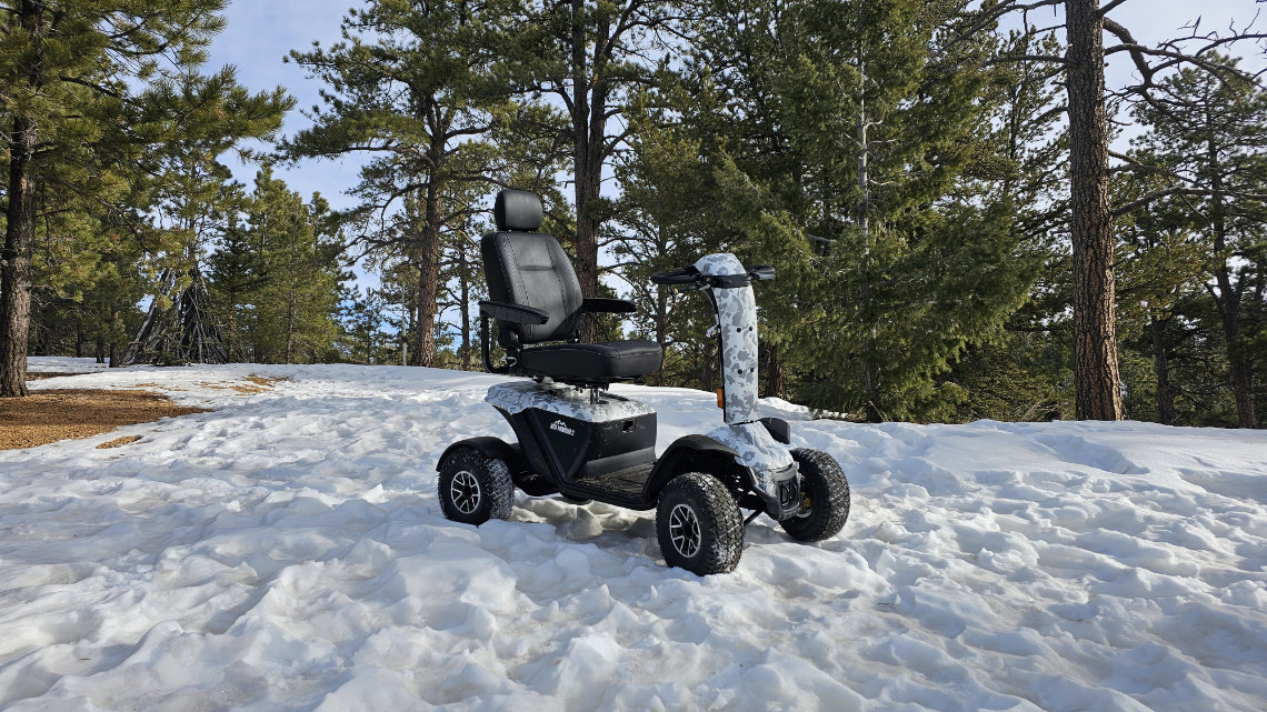 A rugged mobility scooter with large tires is parked on a snowy forest trail surrounded by pine trees.