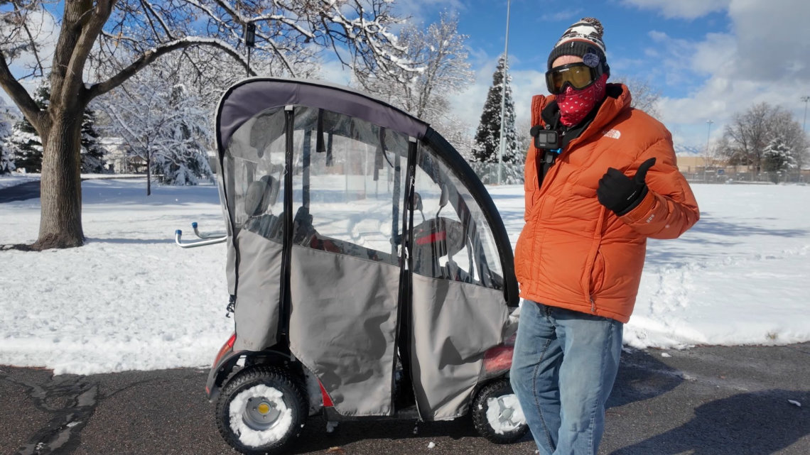 Person in winter gear stands next to a covered mobility scooter in a snowy park, giving a thumbs up.
