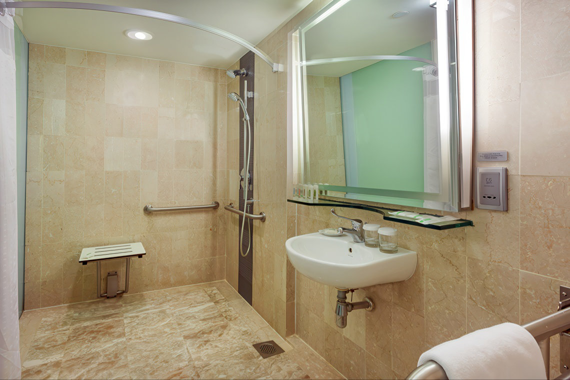 Accessible room bathroom at Holiday Inn Singapore Orchard City Centre with a walk-in shower, fold-down seat, grab bars, and a sink with toiletries under a lit mirror.
