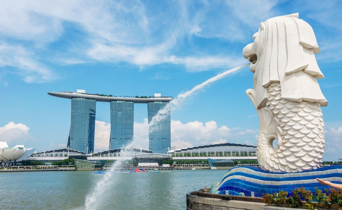 Merlion statue spouting water into Marina Bay with Marina Bay Sands and city skyline in the background under a clear blue sky.