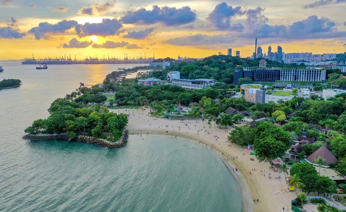 Aerial view of Siloso Beach in Sentosa, Singapore, with lush greenery and sunset over the sea and port cranes in the distance.