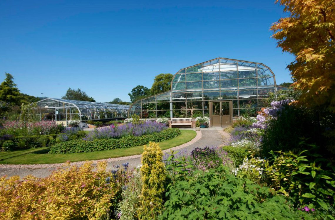 Inverness Botanic Gardens, glass greenhouses surrounded by vibrant flower beds under a bright blue summer sky.