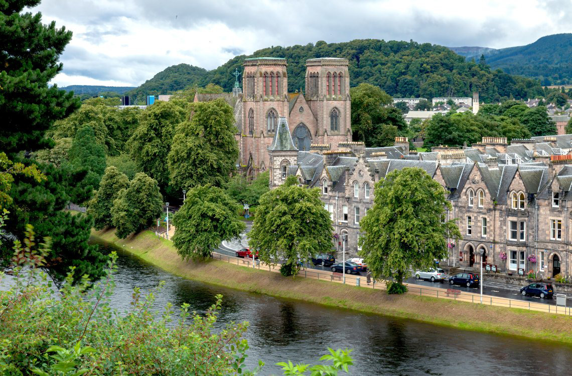 Inverness Cathedral, twin-towered red sandstone church set among trees beside the River Ness with hills in background.