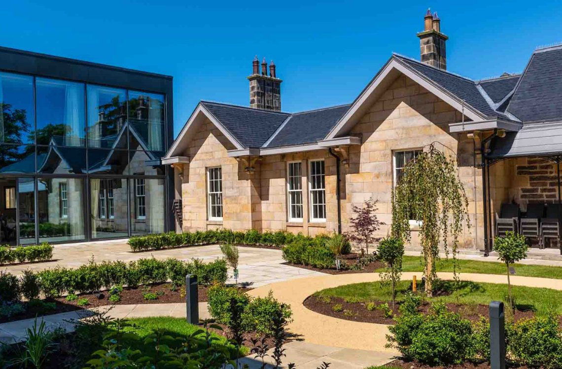 Ness Walk Hotel, a stone-fronted building with gardens, modern glass extension, and clear blue sky.