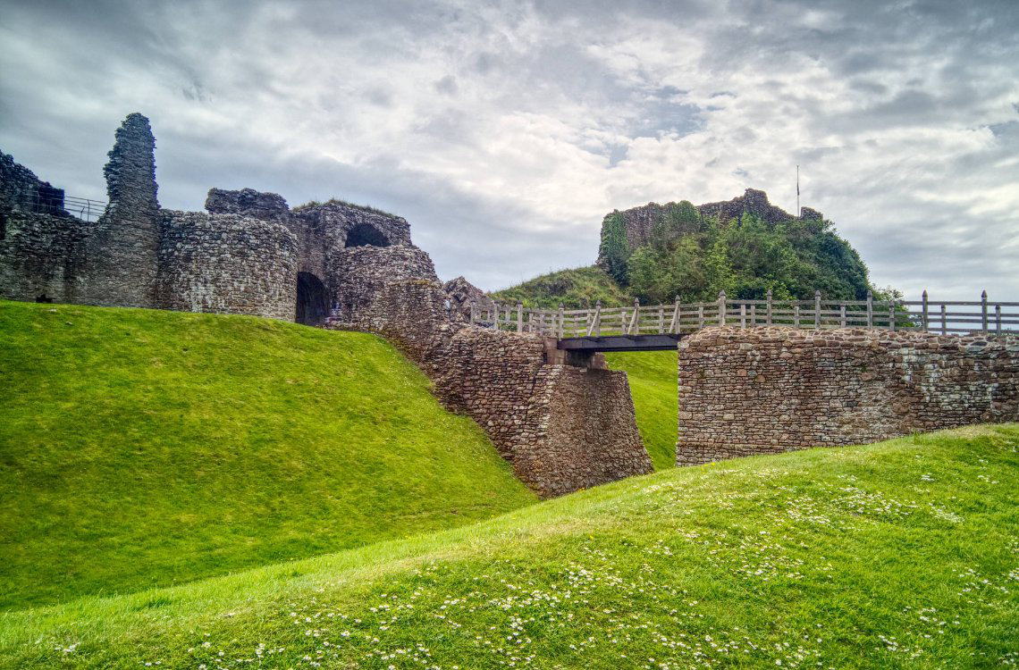Ruins of Inverness Castle, stone walls and bridge over grassy moat under a cloudy sky.